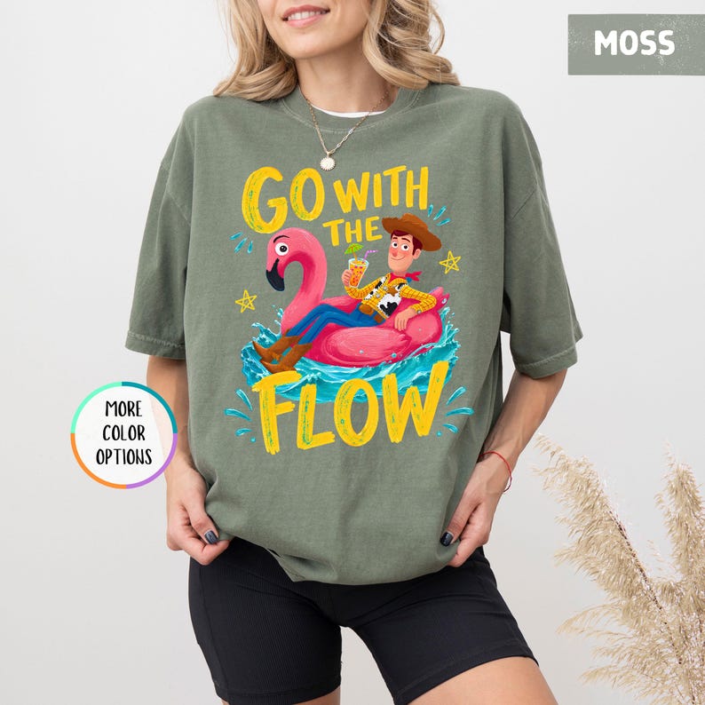 Puede incluir: Una camiseta de manga corta color verde musgo con un gr&aacute;fico de un vaquero sentado en un flotador de flamenco rosa sobre agua azul. El texto "GO WITH THE FLOW" aparece en letras amarillas, junto a peque&ntilde;as estrellas amarillas y salpicaduras.