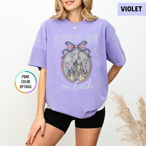 May include: A violet-colored t-shirt with the text "Happiest Mom on Earth" above a castle graphic. The graphic includes a pink bow, balloons, and stars. The shirt has short sleeves and a relaxed fit.