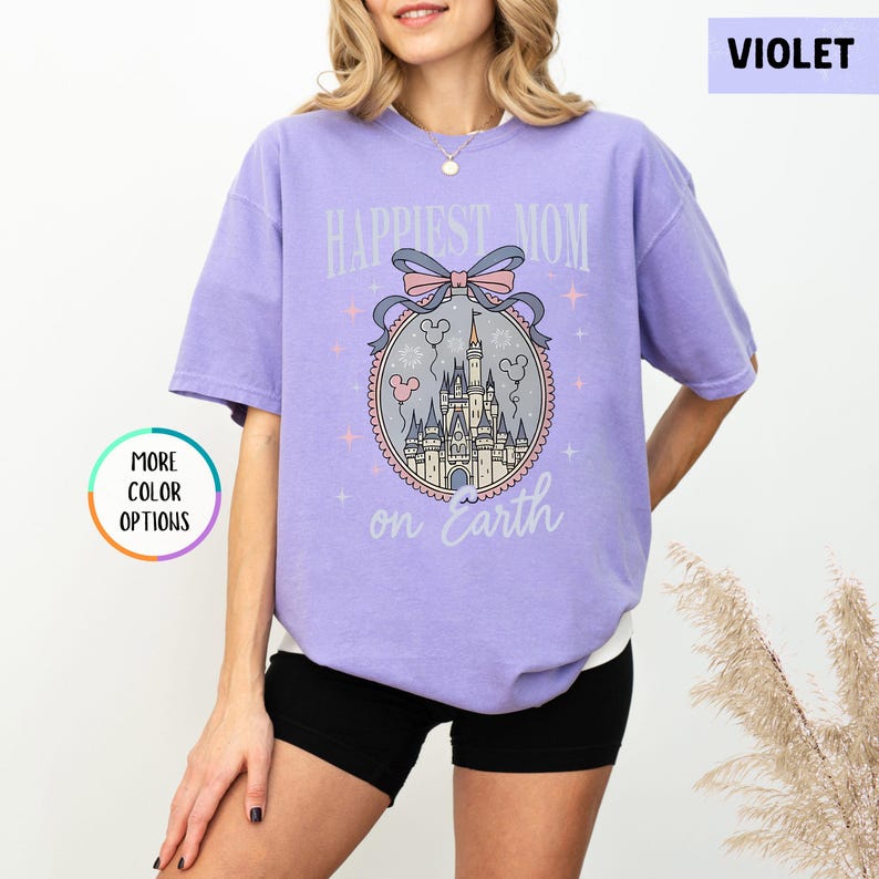 May include: A violet-colored t-shirt with the text "Happiest Mom on Earth" above a castle graphic. The graphic includes a pink bow, balloons, and stars. The shirt has short sleeves and a relaxed fit.