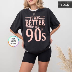 May include: A black short-sleeved t-shirt featuring the text "IT WAS BETTER IN THE 90's" in a distressed light pink font. The shirt has a crew neckline and a loose, relaxed fit. The design is centered on the chest area of the garment.