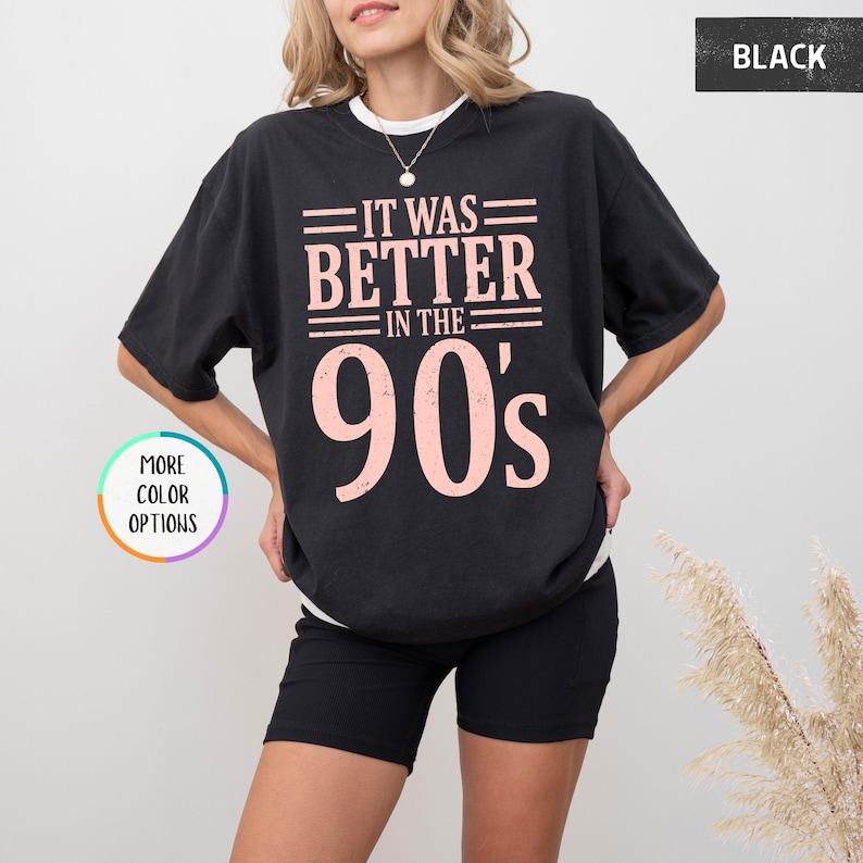 May include: A black short-sleeved t-shirt featuring the text "IT WAS BETTER IN THE 90's" in a distressed light pink font. The shirt has a crew neckline and a loose, relaxed fit. The design is centered on the chest area of the garment.