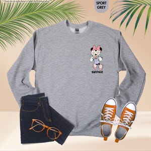 May include: A gray sweatshirt with a Minnie Mouse graphic, the name "SOPHIE" below the graphic, and the text "SPORT GREY" in the upper right corner. The image also shows a pair of dark blue jeans, orange-framed glasses, and orange sneakers.