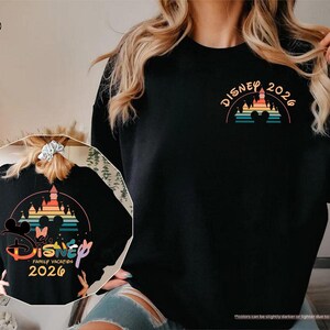 May include: Black sweatshirt with a colorful Disney castle and rainbow design. The front features "Disney 2026" in orange. The back has a larger design with Mickey and Minnie Mouse, the words "Disney Family Vacation 2026".