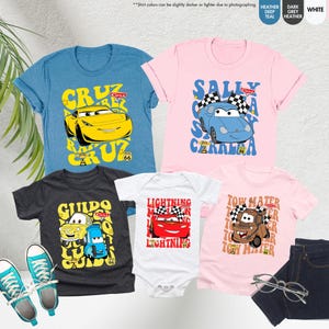 May include: A selection of children's clothing with cartoon car designs. The image shows blue, pink, and grey t-shirts, a white babygrow, and a pair of blue jeans. The shirts feature car character graphics and text, plus a pair of blue trainers.