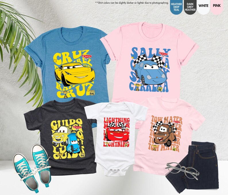 May include: A selection of children's clothing with cartoon car designs. The image shows blue, pink, and grey t-shirts, a white babygrow, and a pair of blue jeans. The shirts feature car character graphics and text, plus a pair of blue trainers.