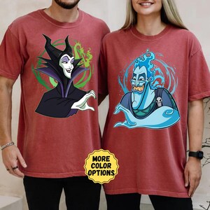 Puede incluir: Dos camisetas granates con personajes de dibujos animados. Una camiseta muestra a Mal&eacute;fica con detalles verdes, la otra a Hades con llamas azules. Las camisetas tienen un corte holgado y el texto "MORE COLOR OPTIONS" es visible.