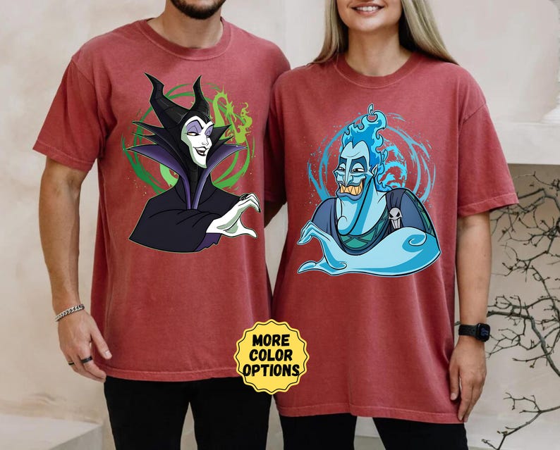 Puede incluir: Dos camisetas granates con personajes de dibujos animados. Una camiseta muestra a Mal&eacute;fica con detalles verdes, la otra a Hades con llamas azules. Las camisetas tienen un corte holgado y el texto "MORE COLOR OPTIONS" es visible.