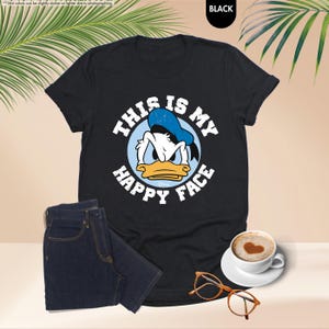 Op de afbeelding: Zwart T-shirt met een afbeelding van Donald Duck en de tekst "THIS IS MY HAPPY FACE". De afbeelding bevat ook een donkerblauwe jeans, een koffiekopje met een hartvorm en een oranje bril.