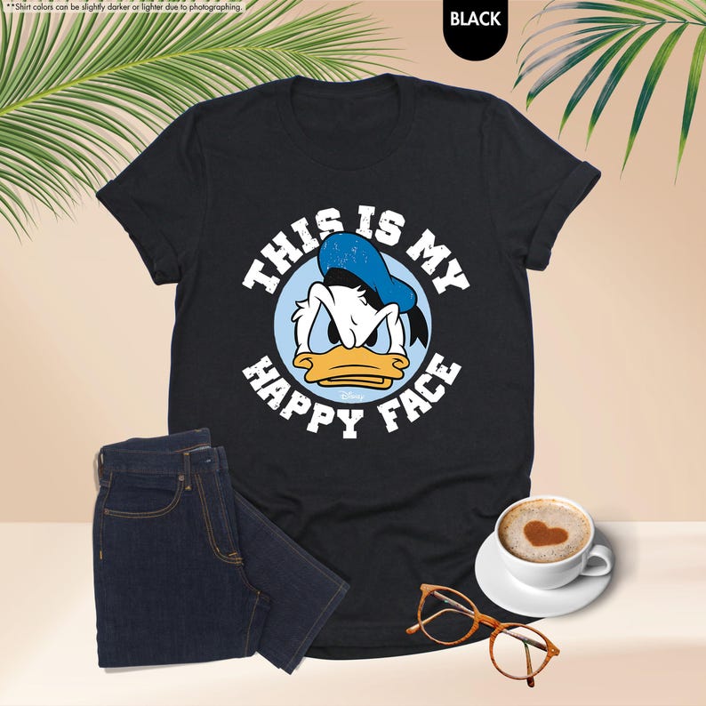 Op de afbeelding: Zwart T-shirt met een afbeelding van Donald Duck en de tekst "THIS IS MY HAPPY FACE". De afbeelding bevat ook een donkerblauwe jeans, een koffiekopje met een hartvorm en een oranje bril.