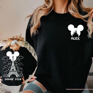 May include: A black long-sleeve sweatshirt featuring a white graphic on the left chest of a Mickey Mouse silhouette containing a castle and the name ALEX. The back design shows a white Eiffel Tower, Mickey ears, clouds, hearts, and DISNEY 2026.