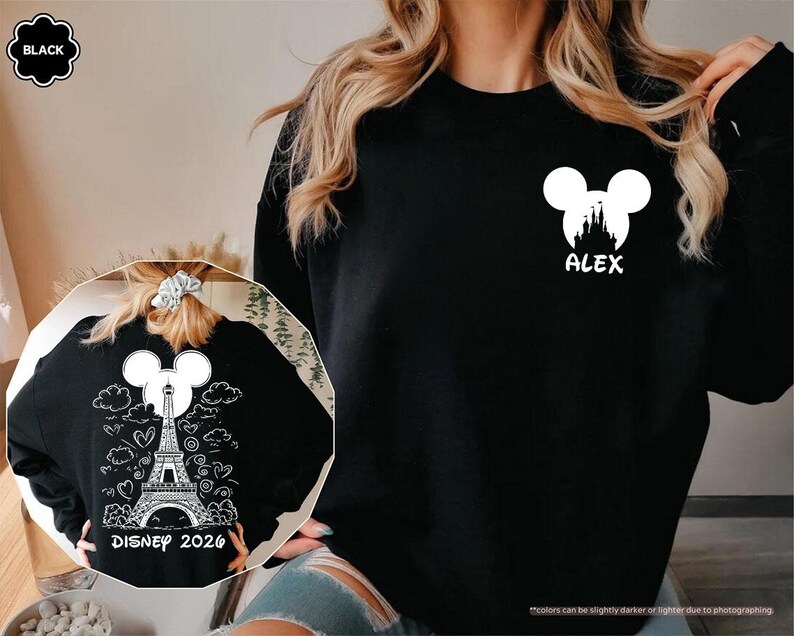 May include: A black long-sleeve sweatshirt featuring a white graphic on the left chest of a Mickey Mouse silhouette containing a castle and the name ALEX. The back design shows a white Eiffel Tower, Mickey ears, clouds, hearts, and DISNEY 2026.
