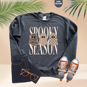May include: A dark heather gray long-sleeve sweatshirt featuring the text SPOOKY SEASON in cream letters. Three ghost illustrations are centered, each with a different pattern: camouflage, leopard print, and a brown and cream checkerboard.