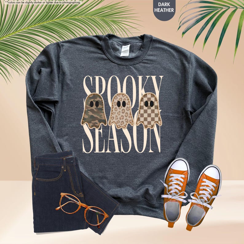May include: A dark heather gray long-sleeve sweatshirt featuring the text SPOOKY SEASON in cream letters. Three ghost illustrations are centered, each with a different pattern: camouflage, leopard print, and a brown and cream checkerboard.