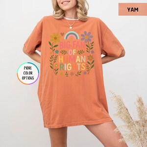May include: A short-sleeved, orange-colored t-shirt with the text "BIG FAN OF HUMAN RIGHTS" in a colorful floral design. The shirt has a relaxed fit and is made of a soft material. The design includes a rainbow and various flowers.