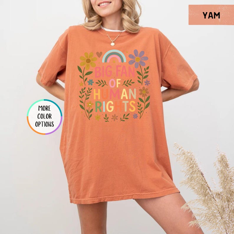 May include: A short-sleeved, orange-colored t-shirt with the text "BIG FAN OF HUMAN RIGHTS" in a colorful floral design. The shirt has a relaxed fit and is made of a soft material. The design includes a rainbow and various flowers.