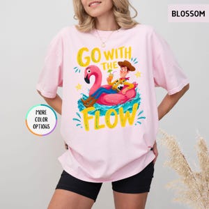 Puede incluir: Una camiseta rosa claro con un gr&aacute;fico de un vaquero sentado sobre un flotador de flamenco rosa en agua azul. El texto GO WITH THE FLOW aparece en letras amarillas. La camiseta tiene mangas cortas, cuello redondo y una etiqueta BLOSSOM.