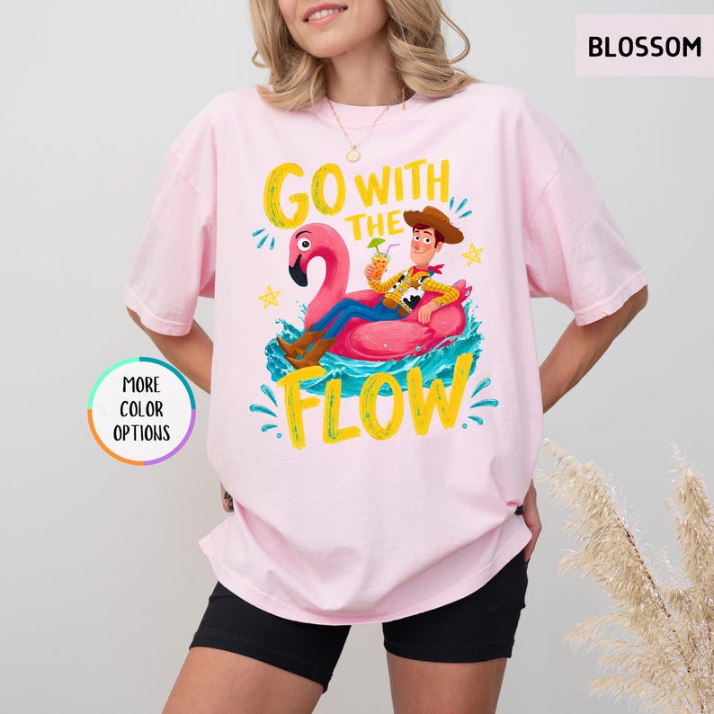 Puede incluir: Una camiseta rosa claro con un gr&aacute;fico de un vaquero sentado sobre un flotador de flamenco rosa en agua azul. El texto GO WITH THE FLOW aparece en letras amarillas. La camiseta tiene mangas cortas, cuello redondo y una etiqueta BLOSSOM.