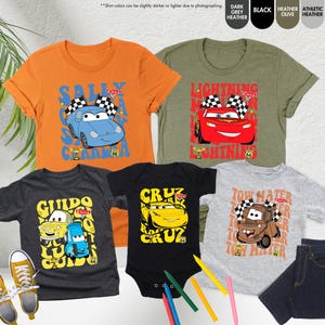 May include: A selection of children's clothing with cartoon car graphics. The image shows orange, olive green, dark grey, and light grey t-shirts, and a black babygrow. The designs feature characters like Sally, Lightning McQueen, Guido, Cruz, and Tow Mater.