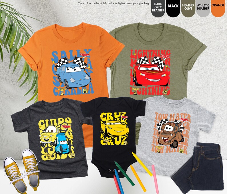 May include: A selection of children's clothing with cartoon car graphics. The image shows orange, olive green, dark grey, and light grey t-shirts, and a black babygrow. The designs feature characters like Sally, Lightning McQueen, Guido, Cruz, and Tow Mater.