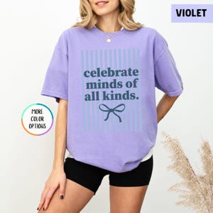 Puede incluir: Camiseta violeta con la frase "celebrate minds of all kinds" en azul oscuro. El texto est&aacute; centrado dentro de un rect&aacute;ngulo a rayas azul claro, con un lazo debajo. La camiseta es de estilo casual.