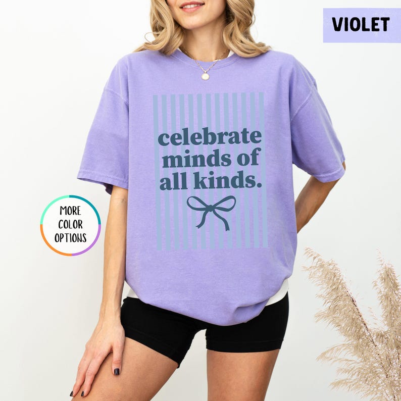 Puede incluir: Camiseta violeta con la frase "celebrate minds of all kinds" en azul oscuro. El texto est&aacute; centrado dentro de un rect&aacute;ngulo a rayas azul claro, con un lazo debajo. La camiseta es de estilo casual.