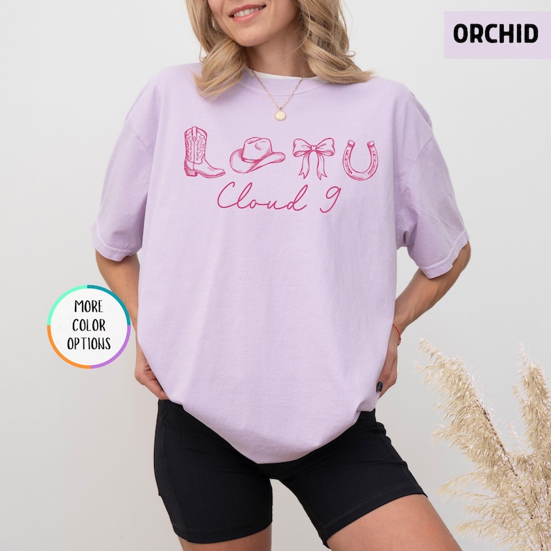 May include: A light purple t-shirt with a pink graphic of a cowboy boot, hat, bow, and horseshoe, with the words "Cloud 9" in a cursive font. The shirt is a solid color and has short sleeves.