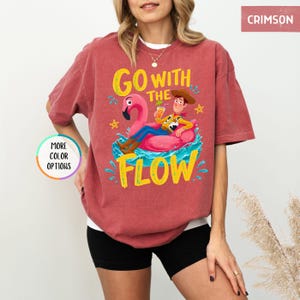 Puede incluir: Una camiseta de manga corta color carmes&iacute; con un gr&aacute;fico de un vaquero sobre un flotador de flamenco rosa en agua azul. El texto GO WITH THE FLOW aparece en letras amarillas. La camiseta incluye una insignia circular con MORE COLOR OPTIONS.