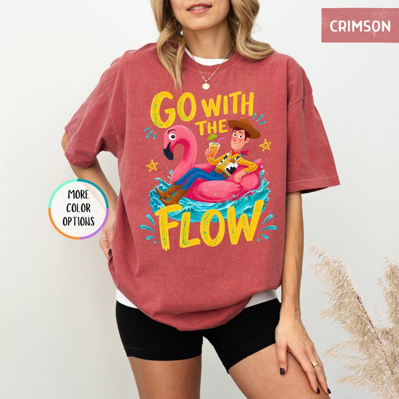 Puede incluir: Una camiseta de manga corta color carmes&iacute; con un gr&aacute;fico de un vaquero sobre un flotador de flamenco rosa en agua azul. El texto GO WITH THE FLOW aparece en letras amarillas. La camiseta incluye una insignia circular con MORE COLOR OPTIONS.