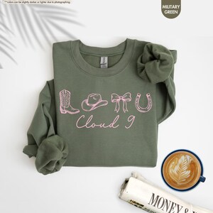 May include: A military green sweatshirt featuring pink cowboy-themed illustrations: a boot, hat, bow, and horseshoe, with the text "Cloud 9". The sweatshirt is on a white surface, with a coffee cup and newspaper in the background.