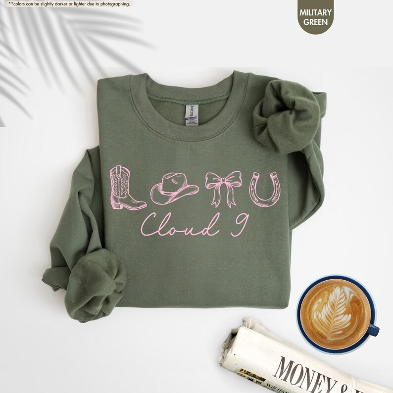 May include: A military green sweatshirt featuring pink cowboy-themed illustrations: a boot, hat, bow, and horseshoe, with the text "Cloud 9". The sweatshirt is on a white surface, with a coffee cup and newspaper in the background.