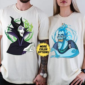 Puede incluir: Camisetas blanquecinas con ilustraciones de dibujos animados de los villanos de Disney Mal&eacute;fica y Hades. Mal&eacute;fica est&aacute; representada en negro y morado, mientras que Hades est&aacute; en azul. Las camisetas tienen cuello redondo y mangas cortas. El texto "MORE COLOR OPTIONS" es visible.