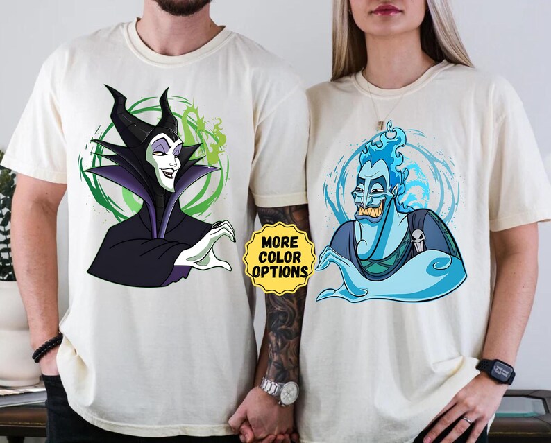 Puede incluir: Camisetas blanquecinas con ilustraciones de dibujos animados de los villanos de Disney Mal&eacute;fica y Hades. Mal&eacute;fica est&aacute; representada en negro y morado, mientras que Hades est&aacute; en azul. Las camisetas tienen cuello redondo y mangas cortas. El texto "MORE COLOR OPTIONS" es visible.