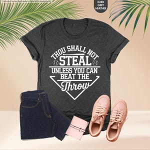 May include: Dark grey heather t-shirt with the text "THOU SHALL NOT STEAL UNLESS YOU CAN BEAT THE THROW" in white. The image also includes a pair of dark blue jeans, a small pink wallet, and a pair of pink sneakers.