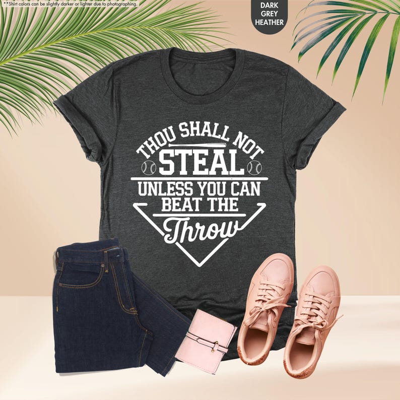 May include: Dark grey heather t-shirt with the text "THOU SHALL NOT STEAL UNLESS YOU CAN BEAT THE THROW" in white. The image also includes a pair of dark blue jeans, a small pink wallet, and a pair of pink sneakers.