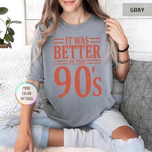 May include: A gray short-sleeved t-shirt featuring the orange text IT WAS BETTER IN THE 90's printed on the front. The shirt has a crew neckline and a relaxed fit. A small circular graphic on the left side displays the text MORE COLOR OPTIONS.