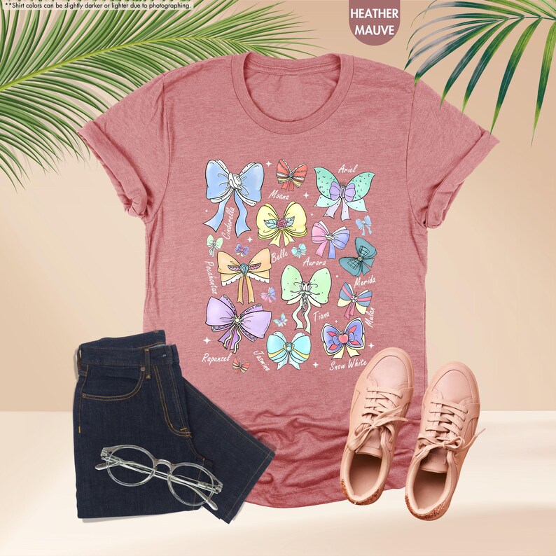 May include: A heather mauve t-shirt with illustrations of colorful bows and character names. The image includes dark blue jeans, clear-framed glasses, and pink trainers. The text "HEATHER MAUVE" is visible.
