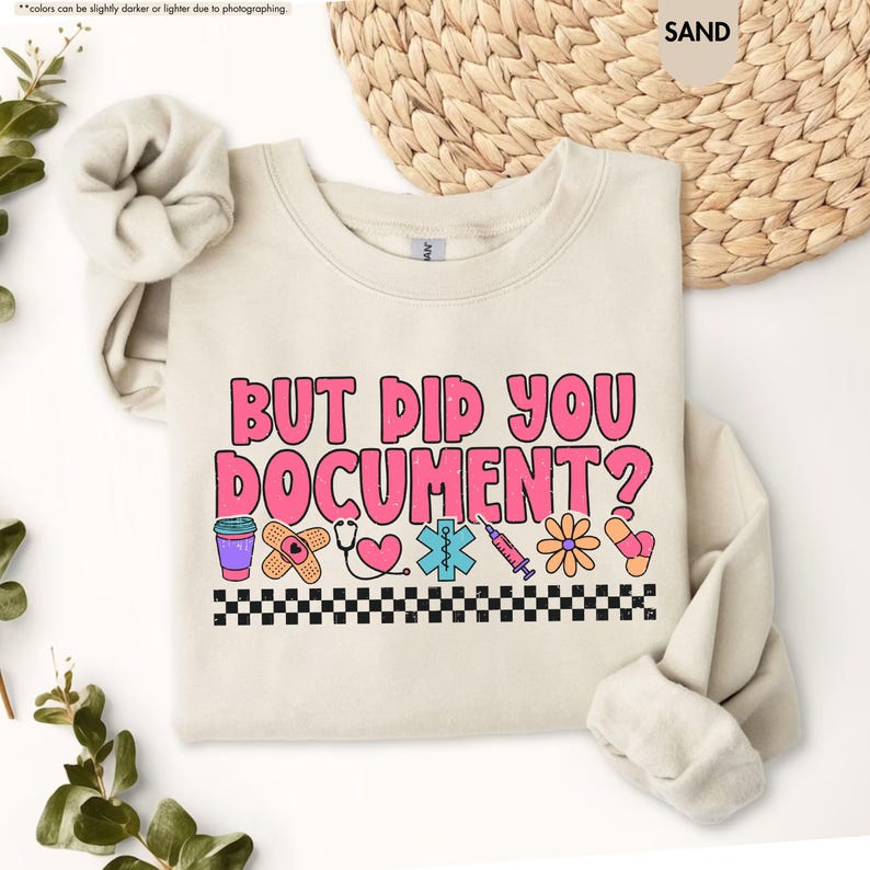 Puede incluir: Sudadera color arena con el texto "BUT DID YOU DOCUMENT?" en rosa. El dise&ntilde;o incluye gr&aacute;ficos m&eacute;dicos como una tirita, un estetoscopio y una jeringa, con un borde a cuadros blanco y negro.