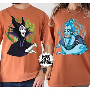 Puede incluir: Dos camisetas de color &oacute;xido con personajes de dibujos animados. Una camiseta muestra a Mal&eacute;fica en negro y morado, con un fondo verde y blanco. La otra camiseta muestra a Hades en azul, con un fondo azul y blanco. El texto "MORE COLOR OPTIONS" es visible.