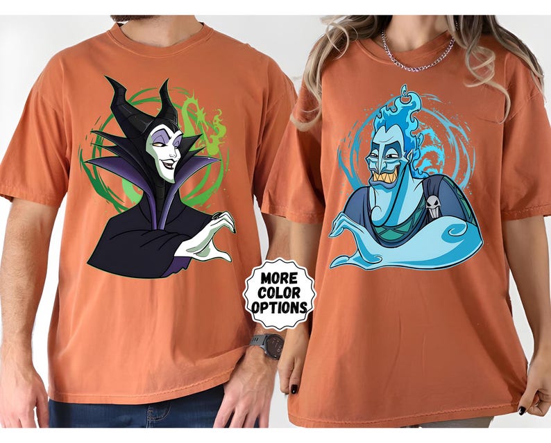 Puede incluir: Dos camisetas de color &oacute;xido con personajes de dibujos animados. Una camiseta muestra a Mal&eacute;fica en negro y morado, con un fondo verde y blanco. La otra camiseta muestra a Hades en azul, con un fondo azul y blanco. El texto "MORE COLOR OPTIONS" es visible.