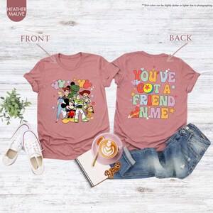 May include: Heather mauve t-shirts with front and back designs. The front features cartoon characters, and the back says "You've Got a Friend in Me." The shirts are displayed with jeans, shoes, and a coffee cup.