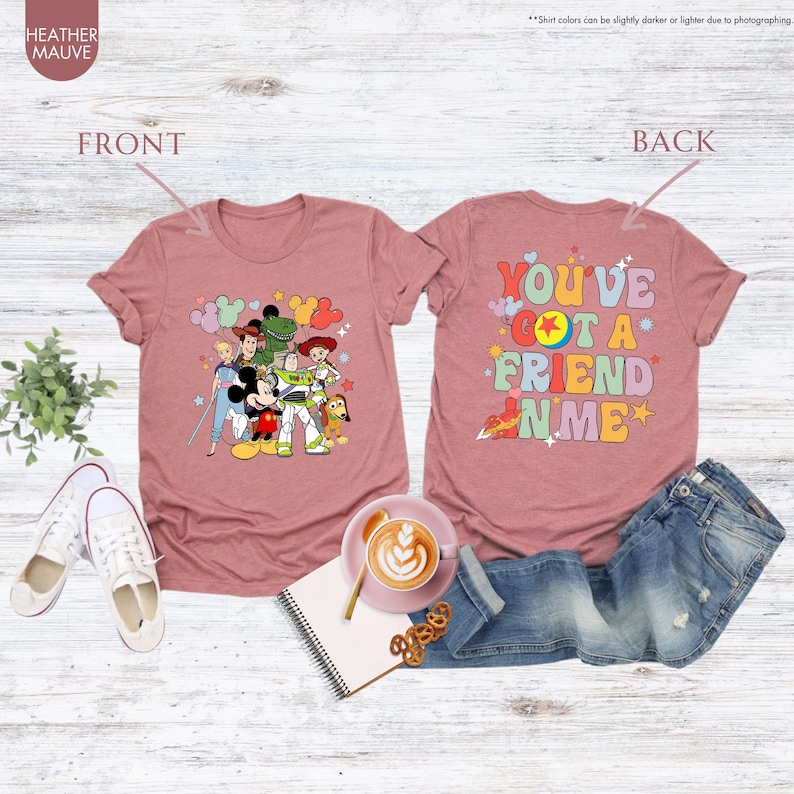 May include: Heather mauve t-shirts with front and back designs. The front features cartoon characters, and the back says "You've Got a Friend in Me." The shirts are displayed with jeans, shoes, and a coffee cup.