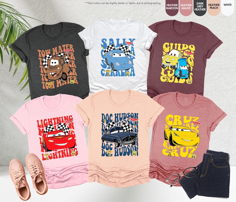 May include: Six t-shirts with cartoon car designs. The shirts are in various colors: dark gray, white, maroon, mauve, peach, and light pink. Each shirt features a different car character with text like 'Tow Mater', 'Sally Carrera', 'Guido', 'Lightning McQueen', 'Doc Hudson', and 'Cruz'.