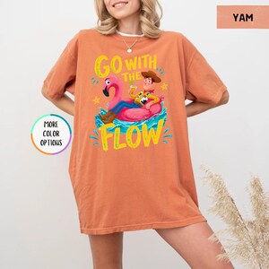 Puede incluir: Una camiseta de manga corta color naranja quemado con un gr&aacute;fico de un vaquero sentado en un flotador de flamenco rosa. El texto GO WITH THE FLOW aparece en letras amarillas. La prenda tiene cuello redondo y una silueta holgada y relajada.