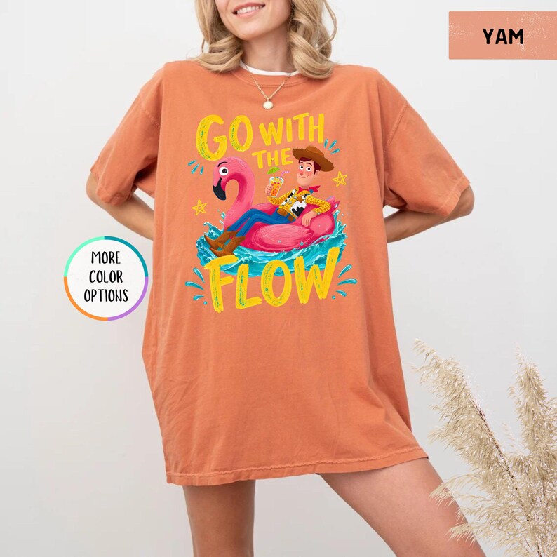 Puede incluir: Una camiseta de manga corta color naranja quemado con un gr&aacute;fico de un vaquero sentado en un flotador de flamenco rosa. El texto GO WITH THE FLOW aparece en letras amarillas. La prenda tiene cuello redondo y una silueta holgada y relajada.
