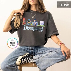 Op de afbeelding: Donkergrijs T-shirt met een Disneyland Resort-afbeelding. De afbeelding toont kleurrijke illustraties van iconische parklandmarks, waaronder een kasteel en een reuzenrad, met het Disneyland-logo in een scriptlettertype. Het shirt heeft een relaxte pasvorm.