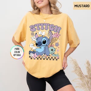 May include: Mustard-colored t-shirt featuring a graphic of Stitch surrounded by flowers and the word "STITCH" in a retro font. The design includes a checkerboard pattern at the bottom. The shirt has a relaxed fit.