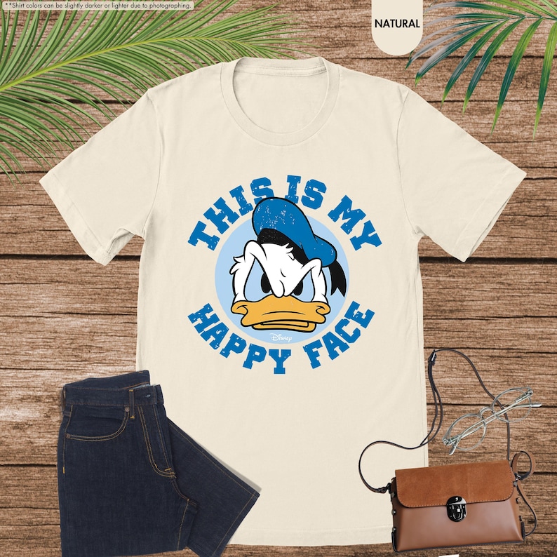 Op de afbeelding: Cr&egrave;mekleurig T-shirt met een afbeelding van Donald Duck en de blauwe tekst "THIS IS MY HAPPY FACE". Het ontwerp toont het gezicht van Donald Duck in een blauwe cirkel. Het shirt wordt getoond met een donkerblauwe jeans en een bruine handtas.