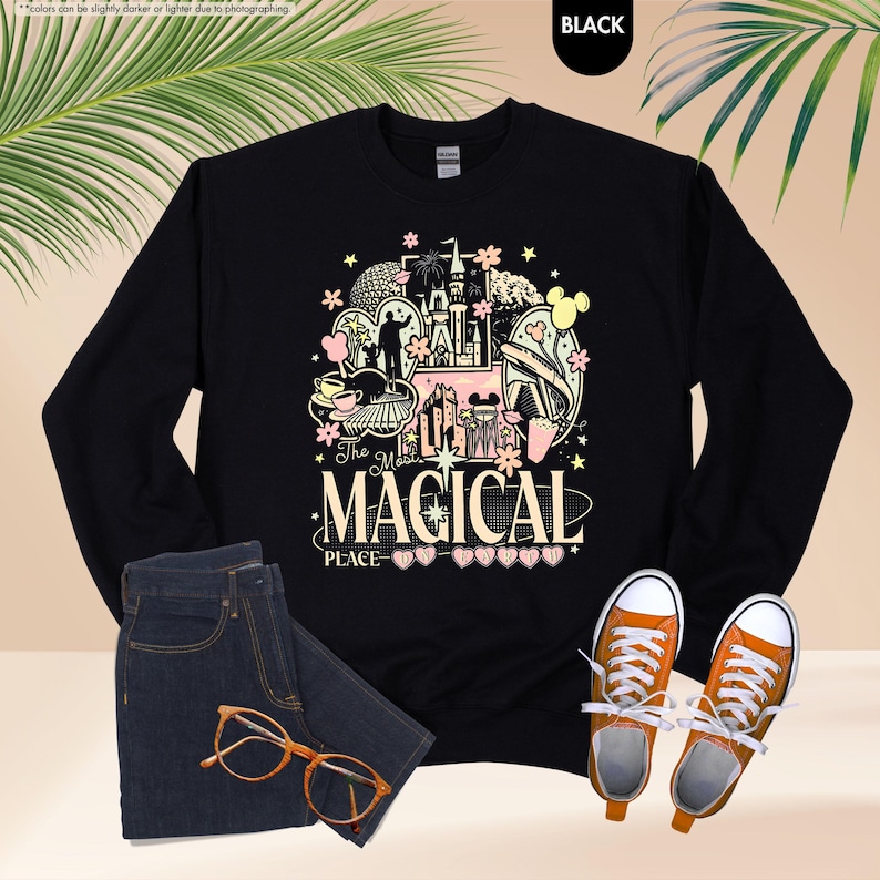 May include: Black long-sleeve sweatshirt with a colorful graphic design featuring a castle, Mickey Mouse, and the text "The Most Magical Place". The sweatshirt is paired with blue jeans, orange glasses, and orange sneakers.