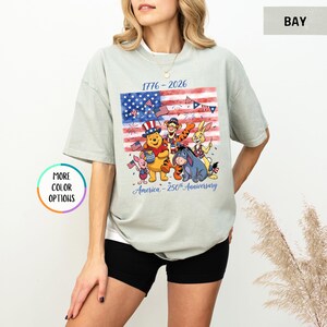 May include: A light green t-shirt featuring a graphic of the American flag and Disney characters. The shirt has the text "1776-2026" and "America - 250th Anniversary". The shirt is a relaxed fit.