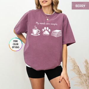 May include: Berry-colored t-shirt with the phrase "My needs are simple." The graphic includes a coffee cup, a paw print, and a stack of books. The shirt is a relaxed fit.